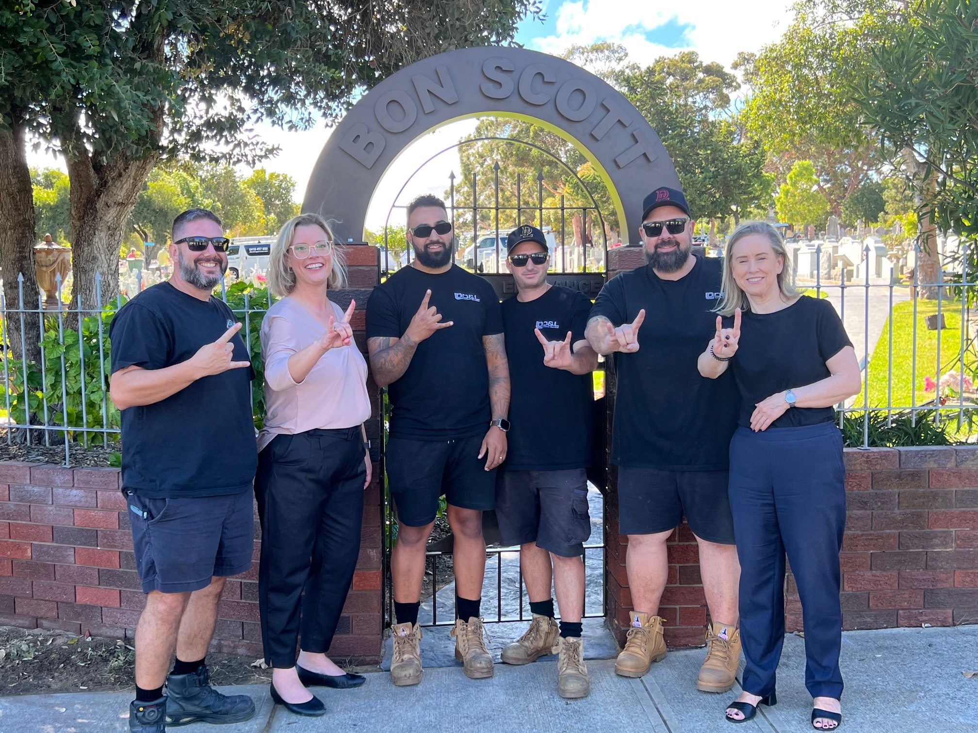 the Hon. Hannah Beazley MLA and Hon. Simone McGurk MLA in the front of the Bon Scott gate at Fremantle Cemetery with the crew of D and L Building Services who restored the gate gesturing the hand sign of 'rock on'